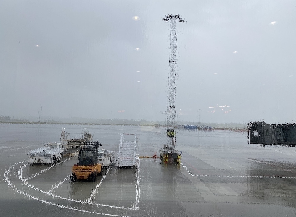 Auto-generated description: A rainy airport tarmac is visible through a wet window, with equipment and a tall light tower in the background.